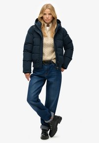 Granatowa kurtka puchowa z kapturem, zapinana na zamek z przodu i przeszyciami. W zestawie z kremowym swetrem w warkocze i ciemnoniebieskimi dżinsami.