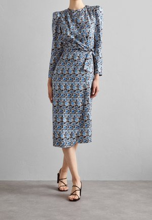 Mujer con vestido midi azul de flores, mangas largas y lazada lateral, combinado con sandalias negras de tacón alto y tiras.