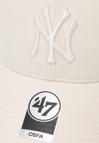 Căciulă de baseball bej cu logo-ul alb în relief al echipei New York Yankees, având un design structurat, detalii cusute și un etichetă de marcă inscripționată '47 OSFA'.