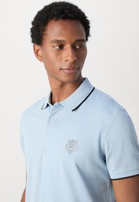 Camisa polo de color azul claro hecha de algodón con un diseño de cuello, que presenta un cuello con borde negro y un emblema de león en el lado izquierdo del pecho.