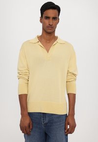 Žlutý pletený svetr s límečkem, dlouhými rukávy a žebrovanými manžetami. Nositý přes tmavě modré džíny s volným střihem.