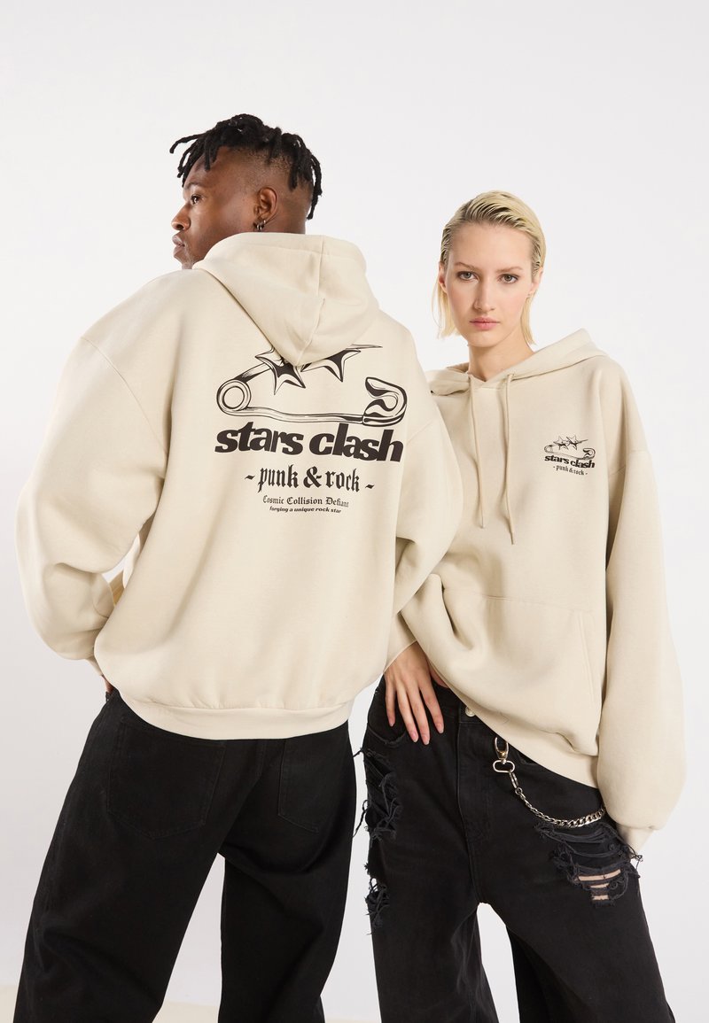 Beige hettegensere med frontlomme, grafiske trykk på bak og bryst, parret med svarte slitte jeans med kjedeaksent.
