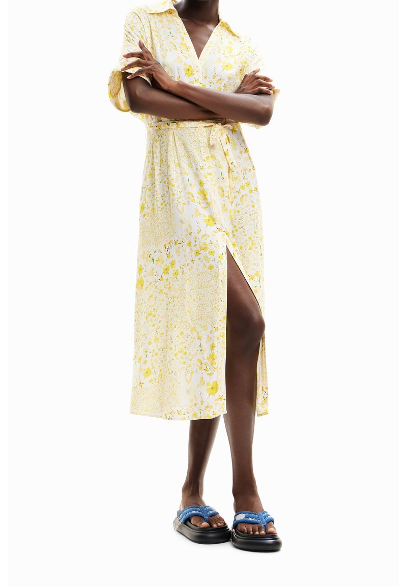 Robe midi florale jaune et blanche avec un col chemise, des manches courtes, une taille ceinturée et une fente sur le côté. Assortie à des sandales bleues.