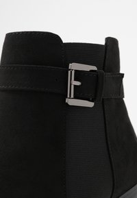 Botín negro con exterior de ante, panel lateral elástico y un acento de hebilla de metal plateado en un diseño suave y afinado.