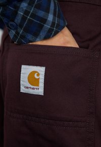 Ranka tamsiai raudonose Carhartt kelnių kišenėje, vilkintis mėlynai juodų kvadratėlių raštą ilgomis rankovėmis marškinėlius. Matomas Carhartt logotipas kišenėje.