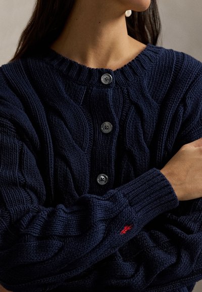 Πλεκτή ζακέτα σε ναυτικό μπλε χρώμα, κατασκευασμένη από υφή υλικό, με έξι στρογγυλά κουμπιά και μικρό κόκκινο κεντημένο λογότυπο στο μανίκι.