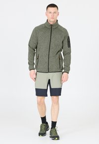 Herrarnas zip-up fleecejacka i mörkgrön, med hög krage och två zip-fickor. Matchas med ljusgrå och marinblå shorts samt svarta skor.