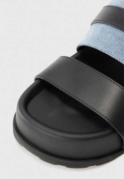 Svarta plattformssandaler med två breda remmar: en i slät svart material och en annan i ljusblå denimtextur.
