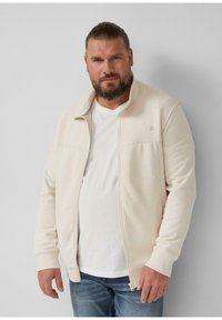 Veste zippée en mélange de coton beige clair avec un col haut, des poignets côtelés et un branding minimaliste sur la poitrine. Portée par-dessus un t-shirt blanc.