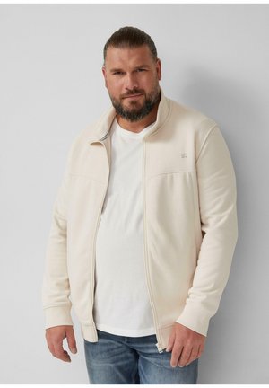 Katoenen blend zip-up jack in lichtbeige met een hoge kraag, ribgebreide manchetten en minimale branding op de borst. Draagt over een wit t-shirt.