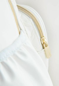 Sac en cuir blanc avec une fermeture éclair dorée et une tirette rectangulaire estampillée de la lettre "B". Le sac présente un design floral texturé.