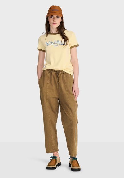 Femme portant un t-shirt graphique jaune, un pantalon marron ample, une casquette beige, des chaussettes rayées et des chaussures marron avec des lacets bleus, debout sur un fond uni.
