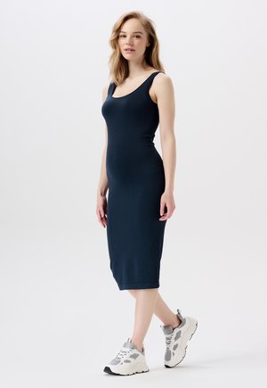 Femme debout portant une robe midi ajustée sans manches bleu marine avec des baskets chunky blanches et grises sur un fond blanc uni.
