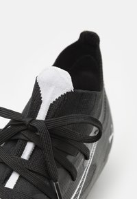Pantof cu aspect sportiv, de culoare neagră, cu o parte superioară din tricot texturat, având accente albe pe vârf și pe șireturi. Șireturile sunt legate într-o fundă.