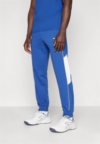 Blue sweatpants with elastic cuffs, featuring white and yellow side panels. Worn with white athletic shoes. Smooth fabric texture.
