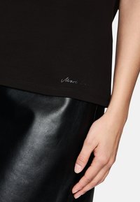 Schwarzes Baumwoll-T-Shirt mit leicht abgerundetem Saum und einem dezenten Marc Cain-Logo am unteren Rand. Kombiniert mit einem glänzenden schwarzen Lederrock.