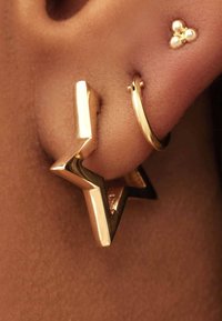 Boucle d'oreille en forme d'étoile en ton or avec une finition polie ; présente un centre creux et un design géométrique. Accompagnée d'un petit stud en or.