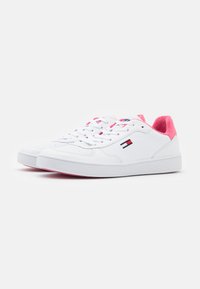 Sneaker sintetiche bianche a taglio basso con accenti rosa sul tallone, lacci bianchi e un sottile dettaglio del logo sul lato. Suola in gomma testurizzata.