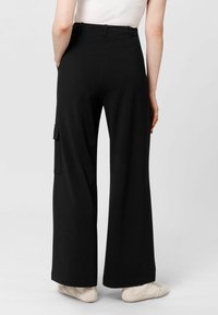 Černé kalhoty s širokými nohavicemi a vysokým pasem, které mají boční kapsu a hladkou texturu látky. Vzhledem k tomu jsou kombinovány se světlými botami.