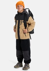 Giacca in pile marrone e nera con zip frontale, abbinata a pantaloni in pile neri. Berretto lavorato a maglia arancione e scarpe da ginnastica grigie. Zaino nero indossato.