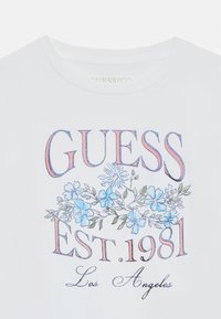 Weißes Baumwoll-T-Shirt mit "GUESS" in blauer und rosafarbener Schrift, floralen Motiven mit hellblauen Blumen und dem Text "EST. 1981 Los Angeles".