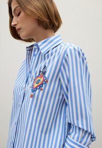 Donna con capelli corti castano chiaro che indossa una camicia a righe blu e bianca con ricami a forma di cuore colorati sul petto.