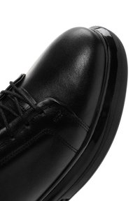 Zapato de cuero negro con un acabado suave, que presenta una punta redondeada, cordones y un acento negro brillante alrededor de la suela.