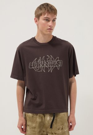 Jonge man met kort lichtbruin haar, die een donkerbruin grafisch T-shirt en lichtbruine gestructureerde broek draagt, staand tegen een effen achtergrond.