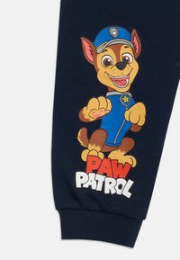 Χαμογελαστός καρτουνίστικος σκύλος που φοράει μπλε αστυνομικό καπέλο και στολή, χτυπώντας με το πόδι πάνω από το κόκκινο και λευκό κείμενο "PAW PATROL" σε ναυτικό ύφασμα.