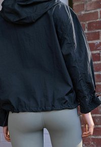 Giacca nera con cappuccio e finitura texturizzata, stretta in vita e ai polsi, abbinata a leggings aderenti grigi chiari.