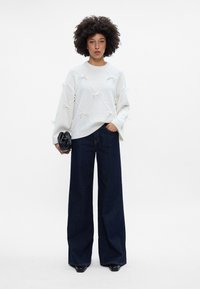 Baltas tekstūruotas megztinis su kaspino akcentais, derinamas su tamsiai mėlynomis plačiomis kelnėmis. Modelis laiko juodą rankinę ir stovi ant vientiso fono.