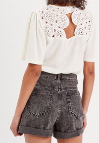 Haut blanc avec un dos en dentelle crochetée, présentant des motifs circulaires et une ouverture ajourée. Associé à un short en denim noir taille haute aux bords roulés.