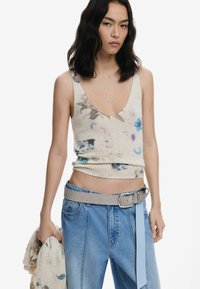 Débardeur en tricot sans manches avec imprimé floral en crème, présentant un décolleté en V profond et une coupe ajustée. Associé à un jean en denim bleu clair et une ceinture grise.