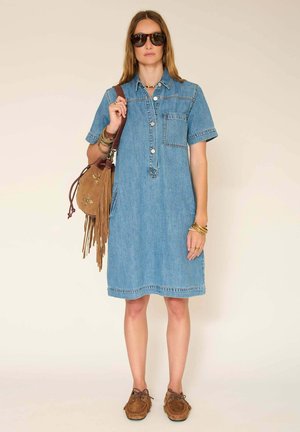 Femme portant une robe en jean bleu jusqu'aux genoux, de grandes lunettes de soleil, des mocassins marron, et un sac à bandoulière marron à franges avec une broderie florale.