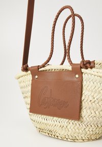 Borsa in paglia intrecciata con una patch in pelle marrone piatta che presenta testo perforato. Manici intrecciati marroni e texture contrastanti.