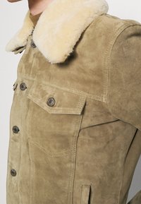 Semišová béžová bunda s měkkým krémovým límcem z ovčí kožešiny, s kovovými knoflíky, dvěma předními kapsami a strukturovaným střihem.