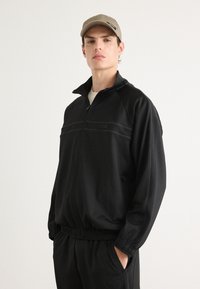 Jaqueta de pista preta com fecho de correr, punhos e bainha canelados. Apresenta costuras contrastantes em cinza e um bolso lateral. Combinada com um boné bege.