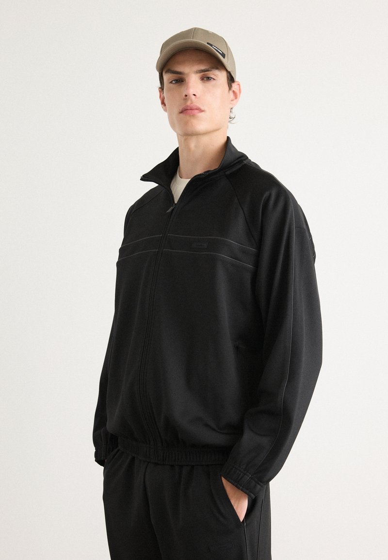 Jaqueta de pista preta com fecho de correr, punhos e bainha canelados. Apresenta costuras contrastantes em cinza e um bolso lateral. Combinada com um boné bege.