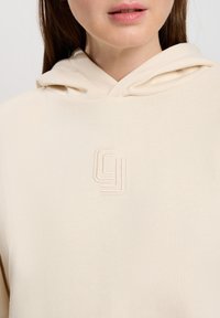 Beżowa bluza z kapturem z kieszenią kangurzą, sznurkiem do regulacji i wytłoczonym logo na piersi. Materiał wydaje się miękki i teksturowany.