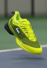 Tennarin kenkä kirkkaassa neon keltaisessa, jossa on teksturoitu päällinen, mustat yksityiskohdat ja kohotettu logo. Varustettu nauhoilla ja pehmustetulla pohjalla.