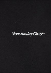 Texte blanc indiquant « Slow Sunday Club™ » sur un fond en tissu noir, probablement une partie d’un vêtement.