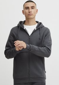 Siva dukserica sa zipom, kapuljačom na str cord, kengur bolso, rebrasti manžeti i rub; meki materijal s suptilnom logom na prsima.