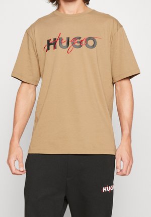 Man wearing beige t-shirt with black and red "HUGO" text and black pants with small matching "HUGO" logo on left thigh.