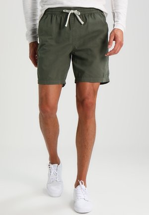 Mann trägt olivgrüne Shorts mit Kordelzug, langärmeliges weißes Hemd und weiße Sneaker, steht vor einem einfarbigen hellen Hintergrund.