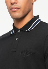 Melnā polo krekls, izgatavots no tekstūras auduma, ar divu pogu aizdari, kontrastējošu jūras zilā un baltā svītru apkakli un iespiestu logotipu uz krūtīm.