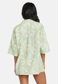 Camicetta verde chiaro a maniche corte con motivo di foglie di palma, tessuto morbido e vestibilità rilassata, con piega sul retro e spacchi laterali.