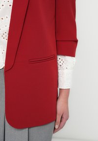 Κόκκινο σακάκι με λεία υφή, διαθέτει πλαϊνή τσέπη και στρογγυλεμένη ποδιά. Κάτω από αυτό, διακρίνεται ένα λευκό κεντημένο πουκάμισο.