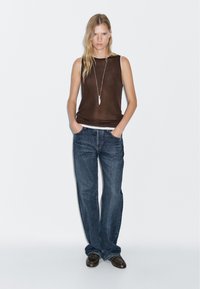 Bruine mouwloze top met een ronde halslijn, gecombineerd met losse donkerblauwe denim jeans. Een lange zilveren hangerketting accentueert de look.