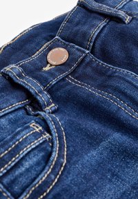 Mørkeblå denimjeans med metalknaplukning, kontrastfarvet syninger og en frontlomme med afrundede kanter.