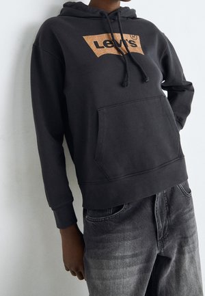 Personne portant un sweat à capuche noir Levi's avec une poche avant et un jean noir délavé, debout devant un fond clair uni.